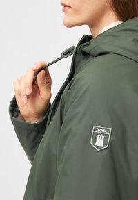 Olivově zelená nepromokavá bunda s kapucí na šňůrku, která má na rukávu logovou nášivku a hladký, lesklý povrch.