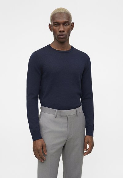 Άνδρας με κοντά ξανθά μαλλιά που φοράει μπλε σκούρο πουκάμισο με μακριά μανίκια και ανοιχτόγκρι παντελόνι ραμμένο στα μέτρα του, με απλό φόντο.