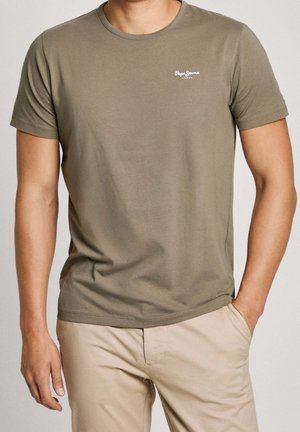 Mand iført olivengrøn kortærmet t-shirt med lille hvidt logo og beige bukser, stående med den ene hånd i lommen.
