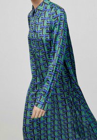 Långärmad skjortklänning i en glänsande tyg med ett geometriskt mönster av blå och gröna former på en mörk bakgrund. Knäppt krage och manschetter.
