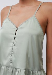 Camisole en satin vert clair avec des fines bretelles, dentelle le long de l'encolure et boutons décoratifs sur le devant. Texture douce et fluide.
