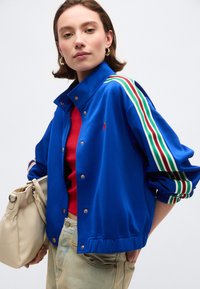 Νεαρή γυναίκα που φοράει μπλε μπουφάν με πράσινες, λευκές και κόκκινες ρίγες στα μανίκια, κόκκινη μπλούζα, ανοιχτόχρωμο τζιν, κρατώντας μπεζ τσάντα, σε λευκό φόντο.