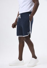 Marineblauwe shorts van zachte stof, met een witte trekkoord, witte zijpiping en een klein logo op de zoom. Gecombineerd met witte sneakers.