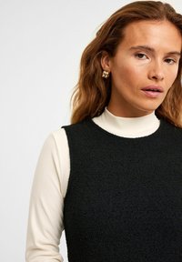 Svart texturerad topp lager på lager över en vit ribbad polotröja. Guldtonade örhängen med ett skulpturalt mönster. Håret i lösa vågor.