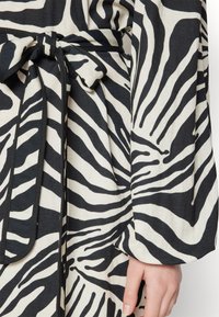 Črno-bela tkanina z vzorcem zebra, ki vključuje pas na zavezovanje, dolge rokave in gladko teksturo, kar poudarja drzen dizajn vzorca.