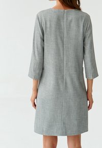Donna che indossa un abito grigio chiaro al ginocchio con maniche a tre quarti e una zip sul retro centrale, in piedi su uno sfondo bianco semplice.