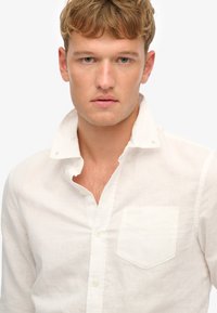 Camisa de lino blanca con collar abotonado y un solo bolsillo en el pecho. La tela tiene un ligero aspecto texturizado.