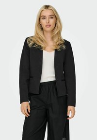 Femme blonde aux cheveux lâchés portant des boucles d'oreilles argentées en forme d'anneau, un blazer noir, un haut blanc et un pantalon noir, se tenant devant un fond uni.