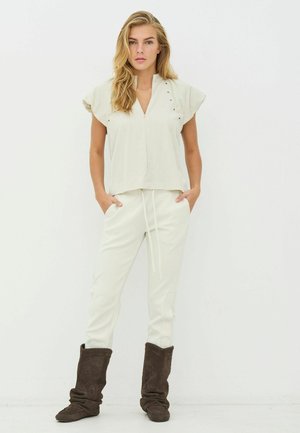 Vrchný diel v krémovej farbe s krátkymi, rozšírenými rukávmi a výstrihom do V, v kombinácii s bielymi nohavicami so sťahovacou šnúrkou a hnedými semišovými čižmami s preloženými manžetami.