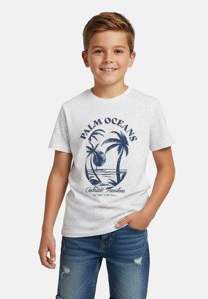 Lachende jongen die een wit "Palm Oceans" T-shirt draagt met palmboomprint en blauwe gescheurde jeans, hand in zak, staand tegen een effen achtergrond.