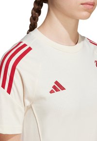 T-shirt en coton crème avec des accents rouges en trois rayures sur les épaules et un logo rouge brodé sur la poitrine. Design classique à col rond.