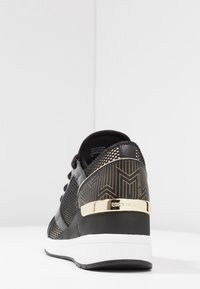 Czarne buty sportowe z teksturowaną siateczką, geometrycznym złotym wzorem na pięcie oraz złotą akcentującą tabliczką z logo. Biała podeszwa.