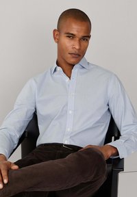 Lichtblauw overhemd met knopen en een subtiel patroon, klassieke kraag en lange mouwen. Bruine korduroy broek met een gladde textuur.