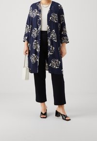 Marineblauer langer Kimono mit weißem Blumenmuster, lockere Passform, 3/4-Ärmel, kombiniert mit einem weißen Oberteil, schwarzen 7/8-Hosen und schwarzen Absatzsandalen.