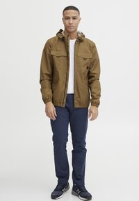 Brown hooded jacket with two front pockets, elastic cuffs, and a grey-lined hood, paired with blue trousers and black sneakers.