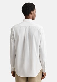 Camicia bianca a maniche lunghe con orlo arrotondato, piega posteriore e polsini con bottoni. Realizzata in tessuto leggero, con colletto classico e texture liscia.