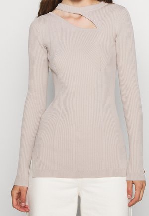 Top beige de manga larga con canalé y un corte asimétrico en el escote, combinado con pantalones blancos, mostrado desde el cuello hasta las caderas.