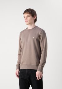 Sudadera de algodón marrón con cuello y puños acanalados, que presenta un pequeño parche de logo en el pecho. El modelo está vistiendo pantalones vaqueros negros.