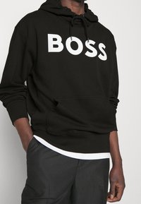 Sudadera negra con capucha y logotipo en blanco "BOSS" en el frente. Tiene un bolsillo tipo canguro y puños acanalados. Debajo, se puede ver el dobladillo de una camiseta blanca.