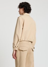 Beige kurze Jacke mit geknöpften Manschetten und Kordelzugsaum. Glatter Stoff, taillierte Passform, minimalistisches Design. Rückansicht.