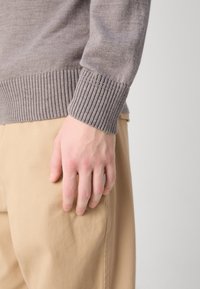 Szürke kötött pulóver bordás mandzsettával, bézs nadrággal viselve. A kéz lazán a lábon nyugszik, megmutatva a bőrszínt és a textúrát.