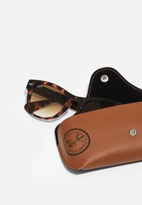 Óculos de sol de tartaruga com lentes castanhas em degradê, apresentando um formato de gato. Acompanhados por uma estojo de couro castanho texturizado, com o logótipo em relevo visível.