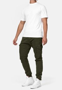 Hombre con camiseta blanca de manga corta, pantalones cargo verdes oscuros con múltiples bolsillos y zapatillas blancas altas contra un fondo blanco.