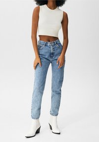 Mujer con un top corto sin mangas blanco, jeans de tiro alto azules y botas de tacón blancas hasta el tobillo, de pie contra un fondo sencillo.