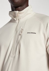 Pullover beige chiaro a zip a quarto realizzato in tessuto testurizzato. Presenta una piccola tasca con zip e un logo stampato sul petto.