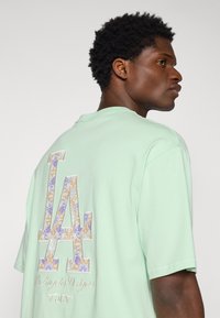 Green cotton t-shirt featuring a large embroidered LA logo in orange and purple floral pattern on the back, with text "Los Angeles Dodgers" underneath.
