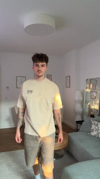 T-shirt oversized bege com um gráfico de planta, combinado com calções cinza claro. O quarto apresenta um chão em madeira clara e uma decoração minimalista.