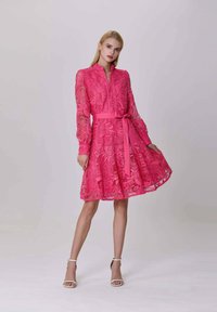 Femme portant une robe en dentelle rose vif à manches longues et à taille nouée, posant avec une main tenant la jupe, chaussée de talons blancs.
