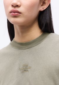 Νεαρό άτομο με μακριά σκούρα μαλλιά φοράει ανοιχτό πράσινο φούτερ κεντημένο με το λογότυπο "Mads Nørgaard Copenhagen".