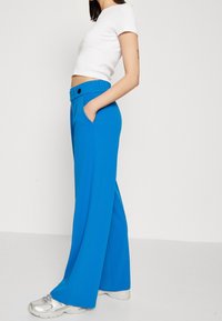 Vida, ljusblå byxor med vida ben och en slät yta, elastisk midja och sidofickor, kombinerade med en figurnära vit topp och silverfärgade sneakers.