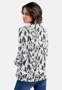 Gestrickte Strickjacke in Schwarz und Weiß mit strukturiertem Blumenmuster, hohem Kragen und lockerem Schnitt. Schulternähte sichtbar.