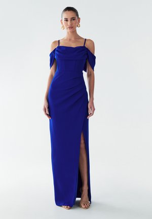 Sieviete, tērpta karaliskā zilo off-plecu kleitā ar plānām siksnām, ar augstu šķēlumu un zelta izteiksmīgajām auskarēm pret vienkāršu fona.