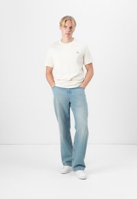 T-shirt en coton blanc avec un col rond, pantalon en denim clair évasé, et baskets blanches, se tenant devant un fond uni.