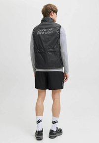 Zwart lichtgewicht vest met hoge kraag, met de tekst "CHASE THE FIRST LIGHT" op de achterkant, gecombineerd met een grijs langarmshirt en shorts.