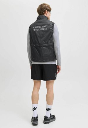 Zwart lichtgewicht vest met hoge kraag, met de tekst "CHASE THE FIRST LIGHT" op de achterkant, gecombineerd met een grijs langarmshirt en shorts.
