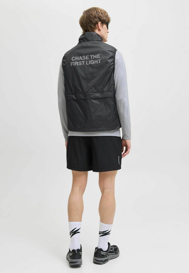 Zwart lichtgewicht vest met hoge kraag, met de tekst "CHASE THE FIRST LIGHT" op de achterkant, gecombineerd met een grijs langarmshirt en shorts.