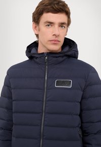 Tamno plava puffer jakna s patentnim zatvaračem sprijeda, kapuljačom i horizontalnim kviltanim uzorkom. Ima logo aplikaciju na prsima i glatku teksturu.