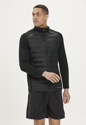 Schwarze Steppweste mit Reißverschluss vorne, taillierter Passform und langen Ärmeln. Getragen über einem schwarzen Langarmshirt und Shorts, mit glatter Textur.