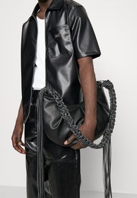 Zwarte leren handtas met een gevlochten handvat en lange franjes. De tas wordt vastgehouden door een persoon die een bijpassende zwarte leren outfit draagt.