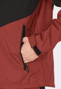 Röd och svart zip-jacka med ficka på sidan, tillverkad av ett slätt, väderbeständigt material. Har passformade ärmslut med kardborreaccenter.