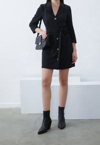 Kvinna som bär en svart klänning med bälte och guldfärgade knappar, svarta ankelstövlar och håller en svart handväska mot en vit och grå bakgrund.