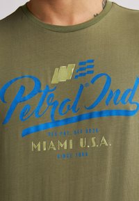 T-shirt vert olive avec des graphismes bleus et jaunes. Présente le logo "Petrol Ind" et le texte "Miami U.S.A. Depuis 1989." Tissu doux et léger.