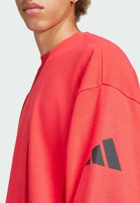 Červená mikina so štruktúrovaným povrchom. Na ľavom rukáve má čierne logo a voľný strih s okrúhlym výstrihom.