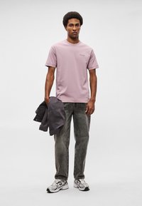 Λιλά βαμβακερό μπλουζάκι με στρογγυλή λαιμόκοψη, συνδυασμένο με γκρι τζιν παντελόνι και σκούρο μπουφάν, φορεμένο με γκρι και μαύρα αθλητικά παπούτσια.