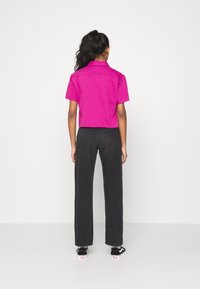 Una camicia fucsia a maniche corte con colletto, abbinata a pantaloni neri dritti e sneakers neri, vista dalla schiena su uno sfondo bianco.