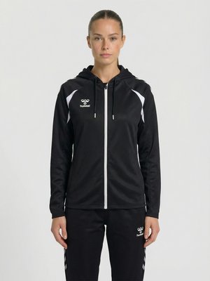 Kvinde iført sort hættejakke med lynlås og matchende sorte bukser med hvidt hummel-logo, stående med front mod kameraet mod en ensfarvet baggrund.